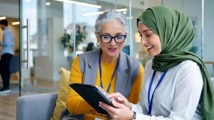 Two diverse businesswomen collaborating and smiling while using a digital tablet in a modern office lounge area. - Powered by Adobe