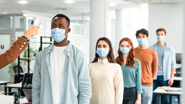 Check Up Measurement. Woman with non-contact laser gun thermometer checking body temperature of multiracial people wearing protective face masks and standing in line at entrance of modern office