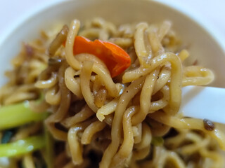 Rolls of springy fried instant noodles are lifted with a fork from the bowl, highlighting their texture and deliciousness, against the blurry background of the bowl.