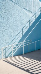 Sunlight creates strong diagonal shadows from a white handrail across a vibrant textured blue wall and a concrete walkway.