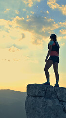 Determined female runner triumphantly stands on a rocky peak at sunset, clad in athletic gear,...