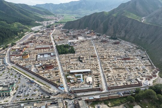 Aerial view of the Labrang Monastery nestled within the town, a tapestry of rooftops and narrow streets against the backdrop of rolling green hills, Xiahe County, China.