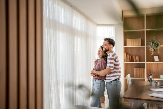 Happy couple embracing and looking out window in new home - Powered by Adobe