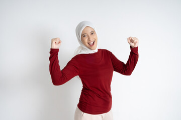 A joyful young Asian Muslim woman wearing a red top and white hijab raises her hands in...