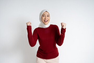 A joyful young Asian Muslim woman wearing a red top and white hijab raises her hands in...