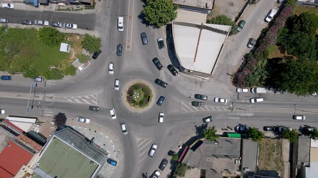 Aerial top-down view of a traffic roundabout in Durr&euml;s, Albania. The intersection features light vehicle traffic, surrounding low-rise buildings, green areas, and a small centered landscaped island.