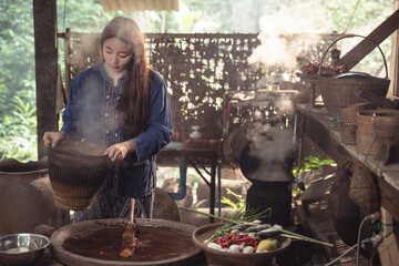A young woman carefully pours hot, steaming sticky rice from a woven basket into a wooden tray. An...