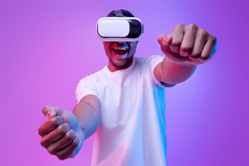 Young african american man playing video games in VR-glasses, excited black millennial guy experiencing virtual reality while standing in neon light over purple gradient background, copy space