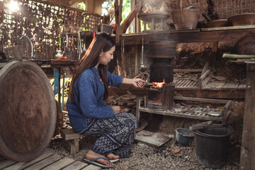 In her rural kitchen, a young woman reaches in to arrange the burning wood in her stove. This wide...