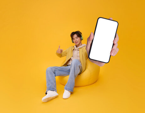 Happy young asian man listening to music with headphone and showing mobile phone sitting bean bag isolated on yellow background. People lifestyle concept. - Powered by Adobe