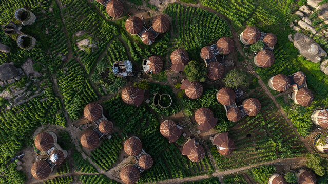 Aerial view of clusters of round, thatched-roof huts nestled amidst lush green fields, creating a textured tapestry from above, Jos, Plateau, Nigeria.
