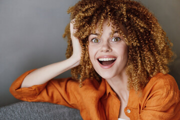 Cheerful young woman with curly hair wearing an orange shirt, expressing joy and excitement in a...