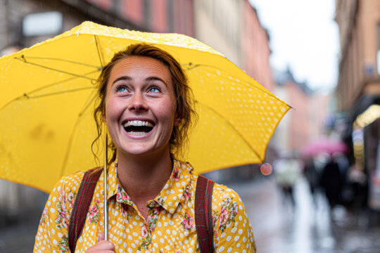 A joyful young woman with curly hair smiles under a yellow umbrella on a city street in rainy weather. - Powered by Adobe