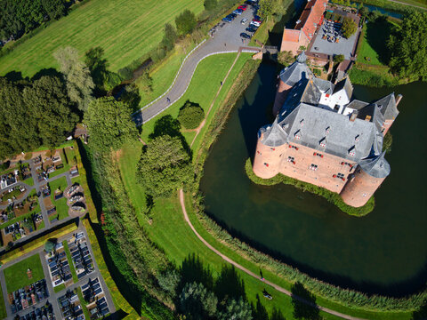 Aerial view of a red brick castle surrounded by a moat and lush green grass, adjacent to a cemetery, Horst aan de Maas, Netherlands.