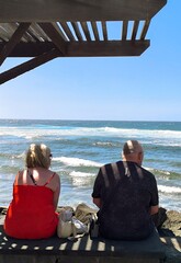 Mature tourist couple sitting and gazing at the sea. Retired people, seen from behind, sitting in front of the sea. Couple sitting and gazing at the horizon, with the sea in the background, on a sunny