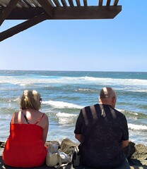 Mature tourist couple sitting and gazing at the sea. Retired people, seen from behind, sitting in front of the sea. Couple sitting and gazing at the horizon, with the sea in the background, on a sunny