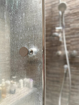 Close-up of a dirty shower glass door with white mineral residue from hard water and soap scum. Blurred shower cabin in the background. Common bathroom cleaning issue.