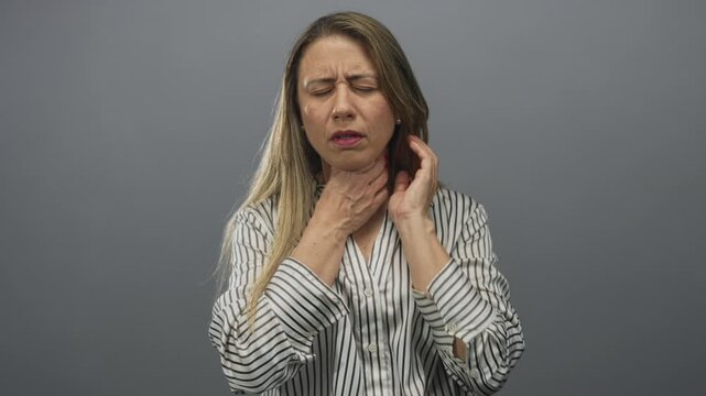 Woman clutches her throat with one hand in gray studio with eyes closed and pained expression; discomfort.