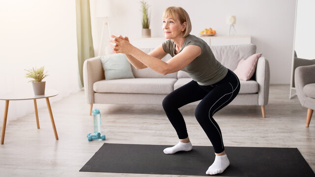 Positive senior lady doing squats on domestic workout in living room, free space. Fit mature woman leading active lifestyle, losing weight, staying in good shape at home during coronavirus quarantine