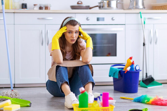 I need break. Exhausted housewife or maid sitting on floor at kitchen among cleaning supplies, holding her head in terror. Tired young woman needing rest from household duties