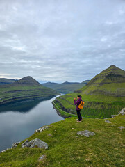 A traveler stands atop a grassy cliff, capturing the stunning view of tranquil waters and lush...