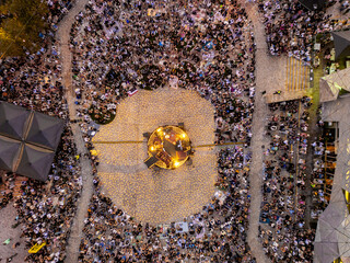 Aerial view of a vibrant crowd gathered around a central stage, bathed in warm light contrasting against the cool evening, Melbourne, Victoria, Australia.