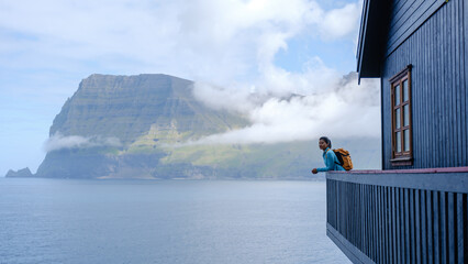 A traveler stands on the edge of a wooden balcony overlooking the dramatic cliffs and calm waters...