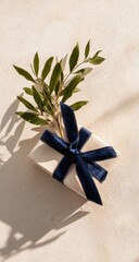 A cream-colored gift box, tied with a navy blue ribbon, and decorated with fresh greenery
