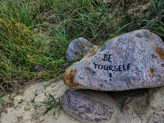 Stone with the inscription 'BE YOURSELF' next to grass and sand in a natural setting. Stone on the shore of the Baltic Sea, 18586 Mönchgut, Reddevitzer Höft, Rügen Island, Germany