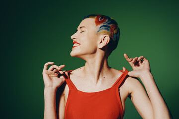 Obraz premium Young woman with colorful shaved hair and red dress smiling joyfully against a green background, expressing happiness and creative self-expression in vibrant style.