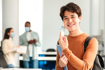 End Of The Epidemic. Smiling asian teen guy student taking off disposable protective face mask,...