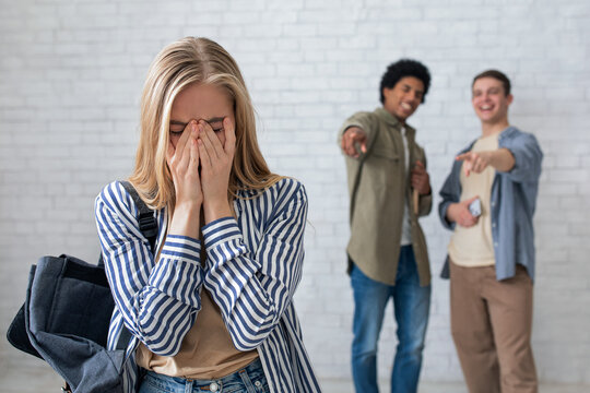 Bullying, negativity and peer problems. European and african american teens zoomers guys laugh at crying female with backpack in school or college, on white wall background, studio shot, empty space