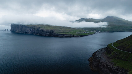 Cloud-laden cliffs rise dramatically above deep blue waters in the Faroe Islands. The tranquil...