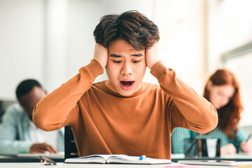 Portrait of shocked asian freshman sitting at table in classroom, grabbing clasping his head in...