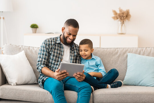 Dad And Son Watching Cartoons On Tablet Online, Sitting Together On Couch Relaxing On Weekend At Home. Happy African American Family Of Two Browsing Internet Using Gadget Computer