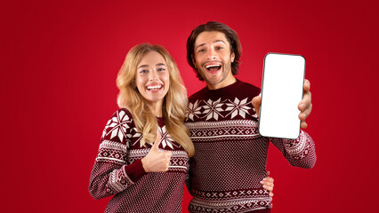 Hapy millennial couple in Christmas sweaters showing thumb up and peace gestures over red background