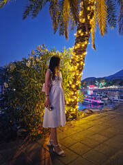 Charming evening scene with a gorgeous woman in a white dress, stands by a palm tree adorned with...