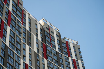 A tall, modern building features a mix of striking red and neutral tones, with numerous large windows reflecting the clear blue sky. The architecture emphasizes a contemporary style