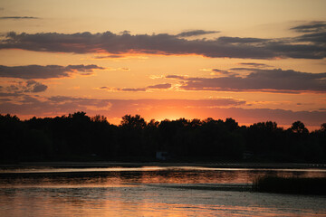 The sun sets over a tranquil lake, casting vivid orange and pink hues across the sky. Silhouettes...
