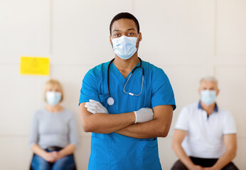 African American male doctor in face mask posing with crossed arms and looking at camera in clinic,...