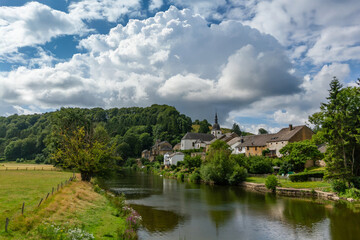 Fototapeta premium Picturesque village nestled by calm river, surrounded by lush greenery and rolling hills. Summer sky with fluffy clouds.