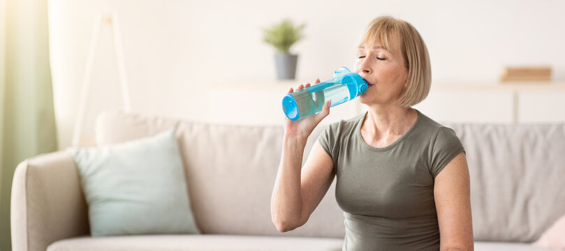 Portrait of fit mature woman drinking water from sports bottle on fitness ball indoors, free space. Active senior lady taking break from domestic training, staying hydrated while working out