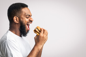 Side View Of Hungry Black Man Eating Burger Posing Standing Over White Background, Studio Shot....