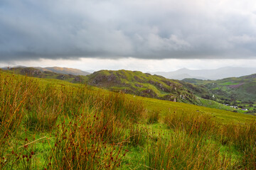 Grassy fields in the foreground lead to green, rocky hills under a cloudy sky in Coomloughra,...