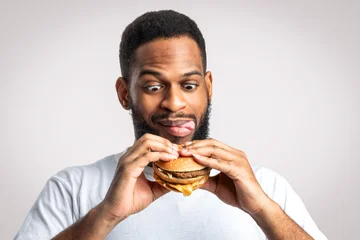 Gordijnen Hungry Black Man Holding Tasty Burger Standing And Licking Lips Posing On White Studio Background. Unhealthy Nutrition And Junk Food Overeating Habit, Cheat Meal Concept © Prostock-studio