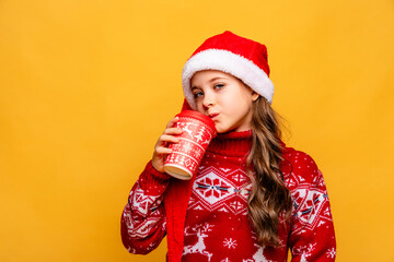 A little girl wearing a red Christmas sweater and a Santa Claus hat drinks coffee or tea from a festive paper cup on yellow background. Happy New Year holiday concept. Copy space. High quality photo