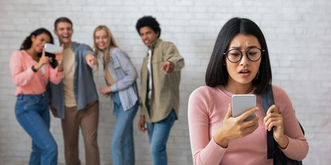 Humiliation, education and bullying. Sad asian young female in glasses looks at phone, zoomers multi ethnic students make video and laugh at lady, on white brick wall, copy space, studio shot