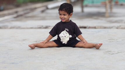 A cheerful 20-month-old toddler sits on the ground outdoors, laughing loudly with a wide smile...