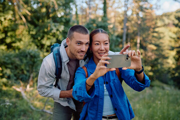 Fototapeta premium Young couple of hikers capturing a joyful selfie with their smartphone while enjoying a beautiful trekking day in the lush forest