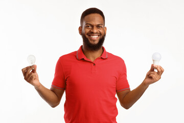 African American Guy Holding Two Light Bulbs Standing On White Studio Background, Choosing...